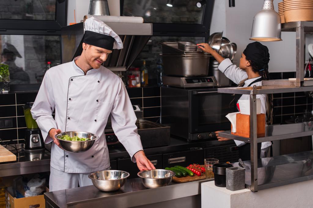 cheerful chef holding bowl with salad near vegetables and african american sous chef in kitchen  - Foto, Imagem