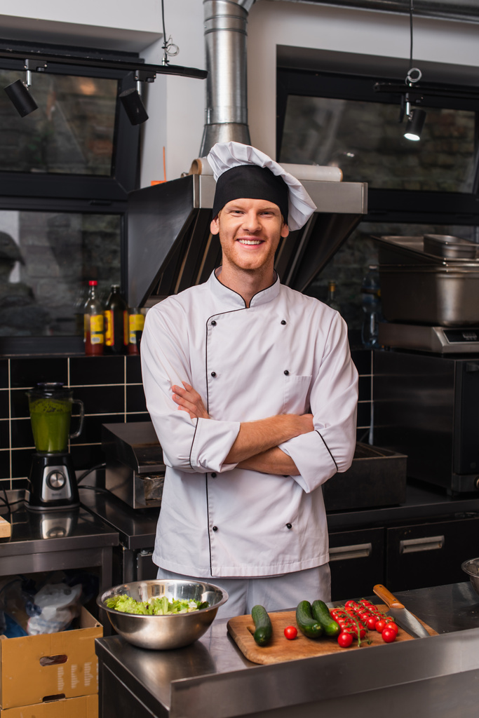 happy young chef in uniform standing with crossed arms near vegetables on cutting board in kitchen  - Zdjęcie, obraz