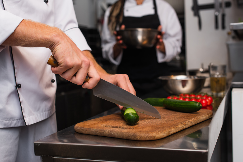 cropped view of chef cutting cucumber on wooden cutting board near blurred colleague  - Zdjęcie, obraz