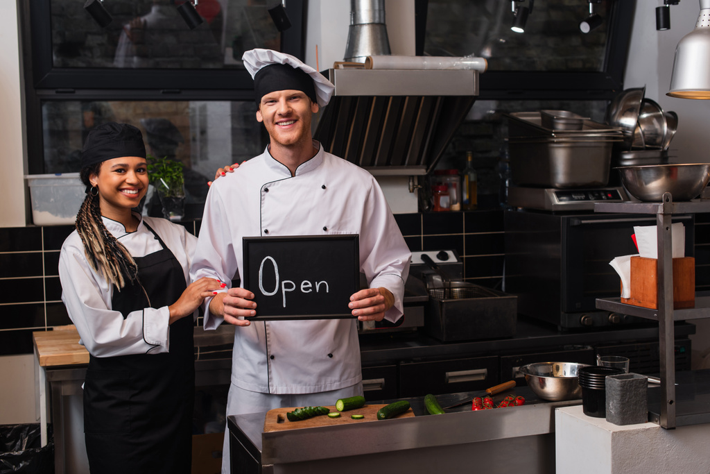 happy young chef in uniform holding chalkboard with open lettering near african american colleague in kitchen  - Photo, Image