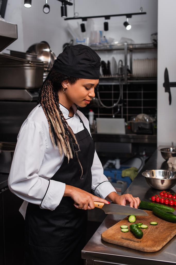 side view of african american woman in apron cutting cucumber in modern kitchen  - Φωτογραφία, εικόνα