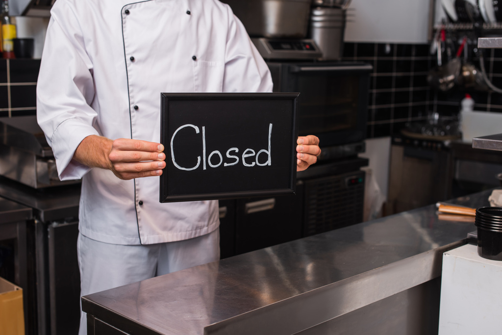 cropped view of chef in uniform holding chalkboard with closed lettering in kitchen - Photo, image