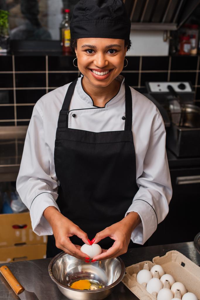 happy african american chef holding raw egg above bowl while cooking in kitchen  - Foto, Imagem