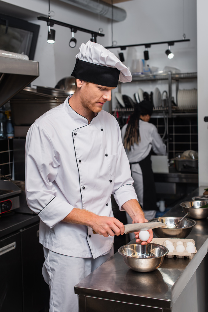 шеф-кухар в уніформі тримає ніж біля сирого яйця над мискою під час приготування на кухні
 - Фото, зображення