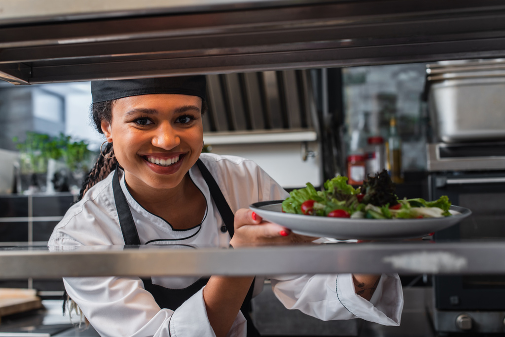 happy african american chef holding plate with freshly cooked salad in professional kitchen - Фото, изображение