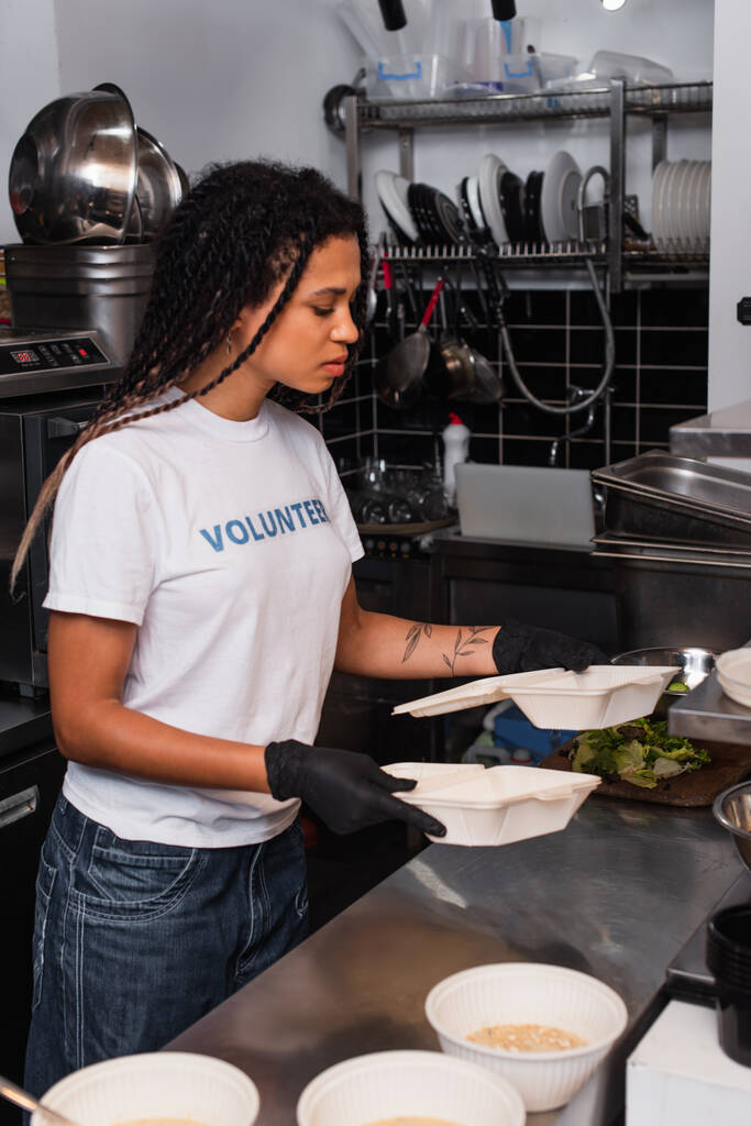 tattooed african american woman in t-shirt with volunteer lettering holding plastic containers in kitchen - Photo, image
