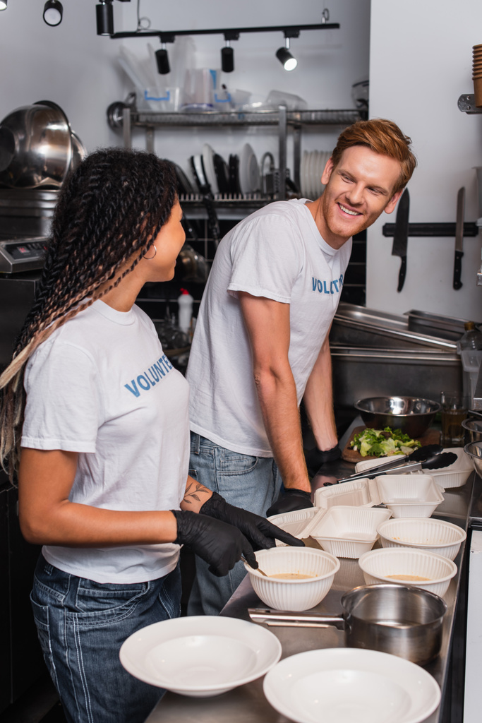 happy interracial volunteers in t-shirts with lettering near plastic containers in kitchen - Photo, image