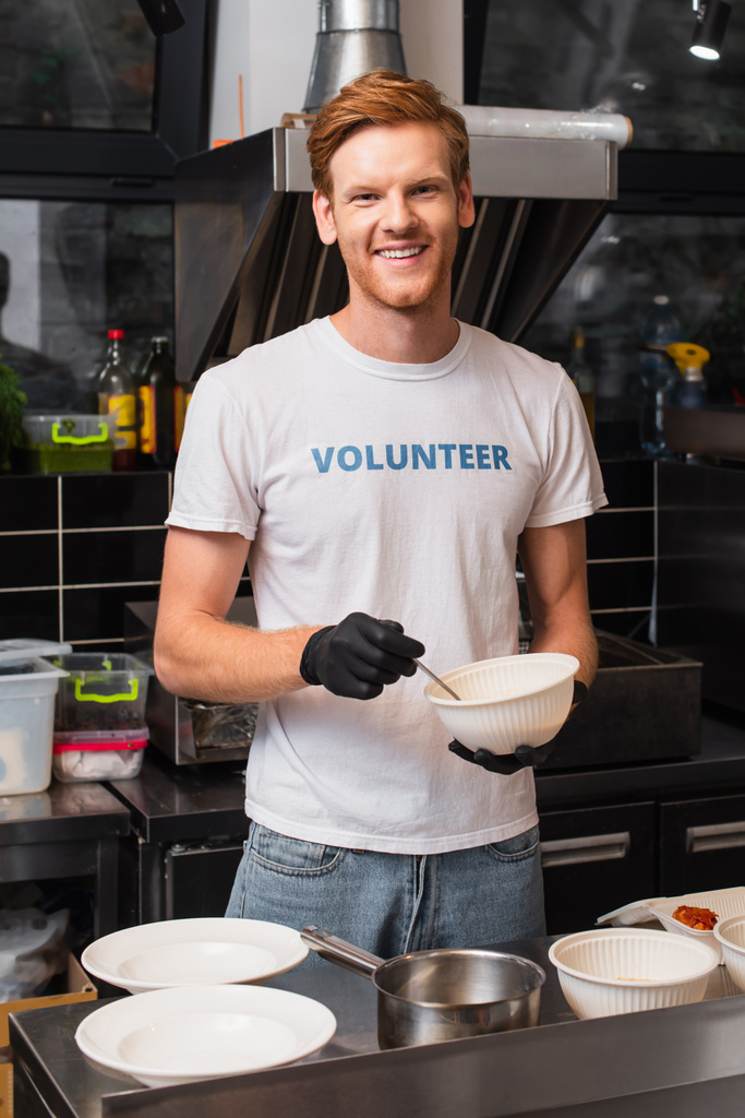 happy and redhead volunteer in latex gloves holding plastic bowl in kitchen  - Valokuva, kuva