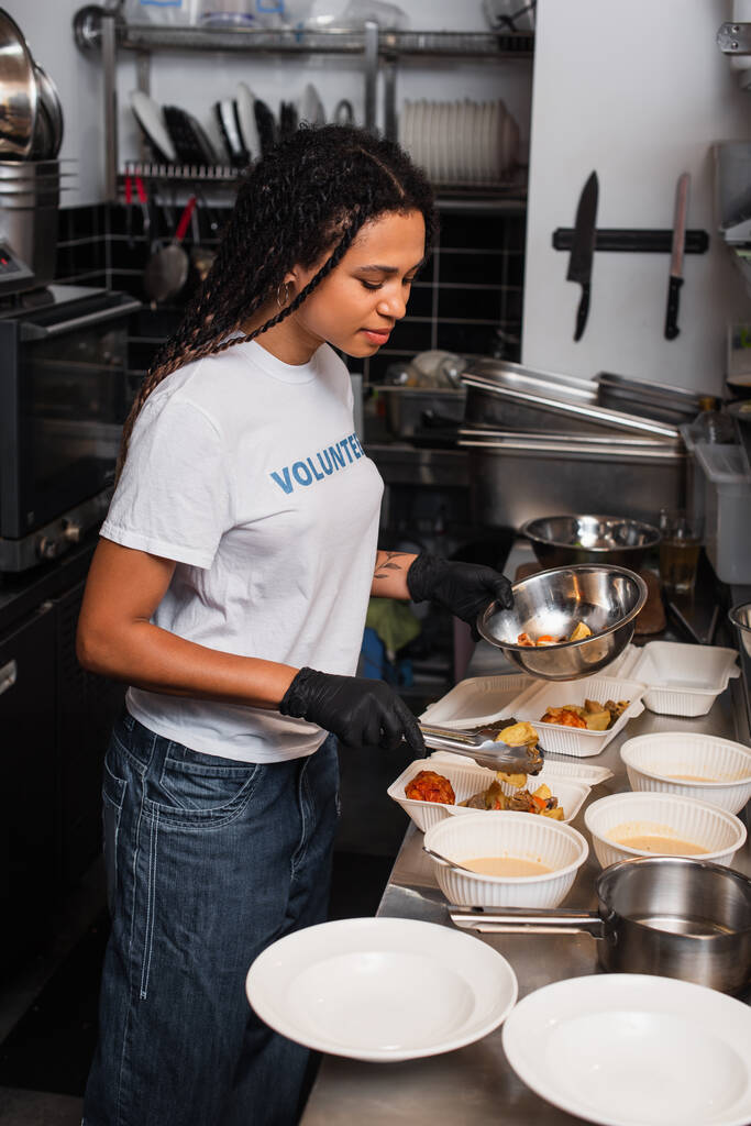 young african american social worker putting prepared meal into plastic containers in kitchen - 写真・画像