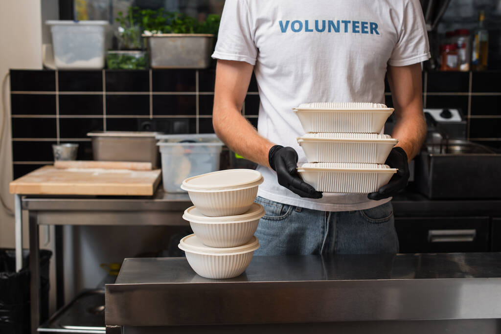 cropped view of man in t-shirt with volunteer lettering holding plastic containers in kitchen  - Foto, afbeelding