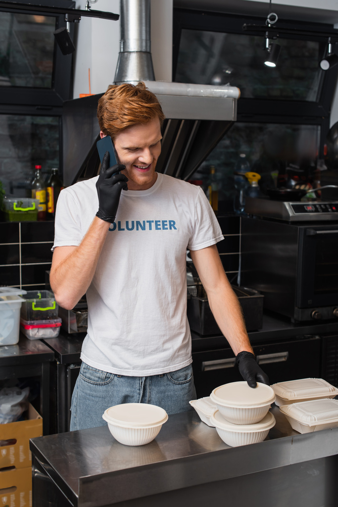 redhead man in t-shirt with volunteer lettering smiling and holding plastic bowl and talking on smartphone in kitchen  - Фото, изображение