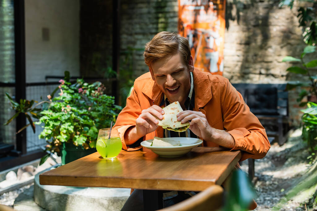 cheerful redhead man eating quesadilla near glass with cocktail in outdoor terrace  - 写真・画像