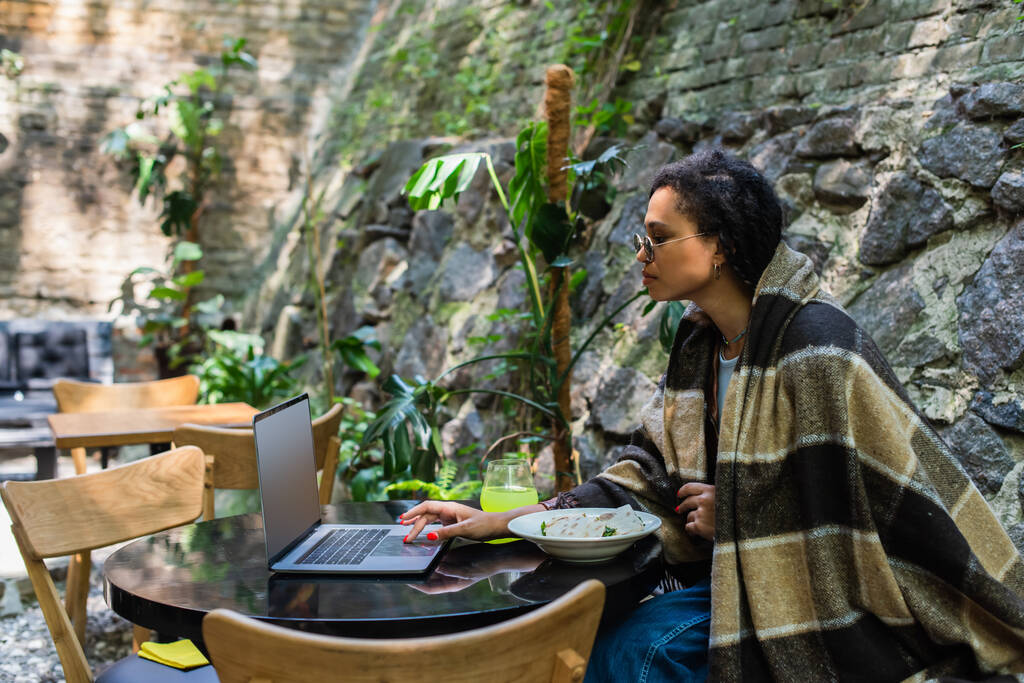 african american freelancer covered in blanket using laptop and having lunch in cafe on terrace - Фото, изображение