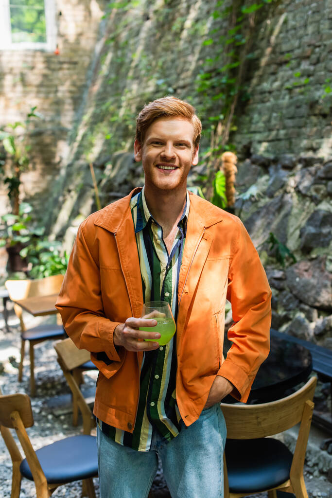 cheerful redhead man in orange jacket standing with hand in pocket and holding glass with cocktail  - Φωτογραφία, εικόνα