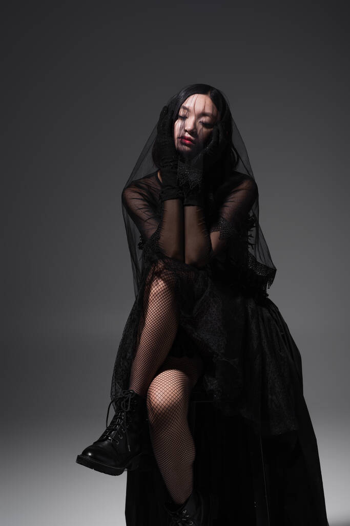 молодая азиатка в готическом наряде с черной вуалью, сидящая на сером - Фото, изображение