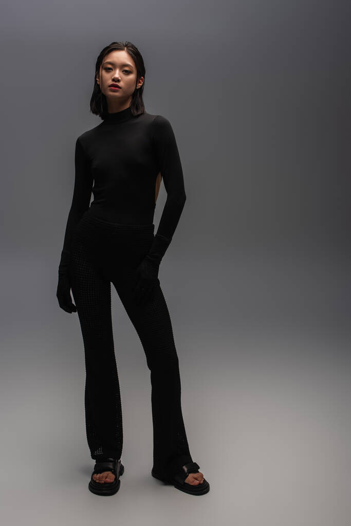 полная длина стильной азиатской женщины в полностью черном наряде, стоящей на сером  - Фото, изображение
