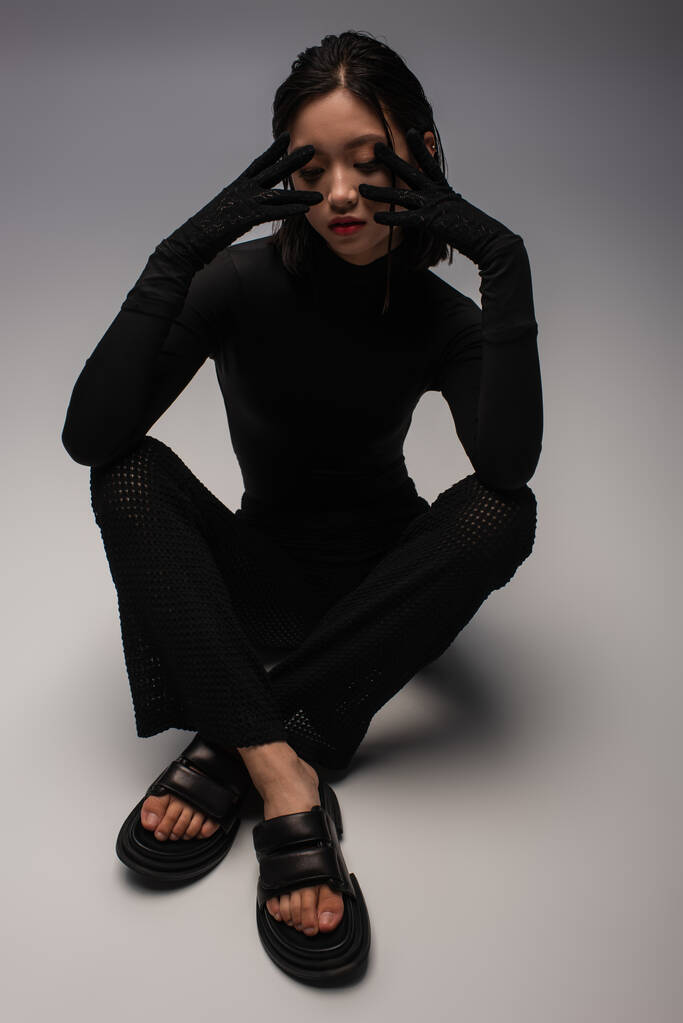 full length of stylish asian woman in total black outfit and gloves touching face on grey  - Foto, Imagen