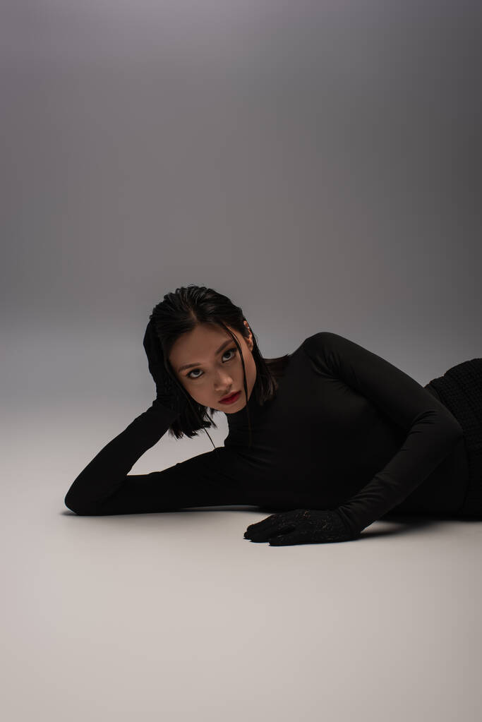 стильна і молода азіатська модель в чорному вбранні лежить на сірому
  - Фото, зображення
