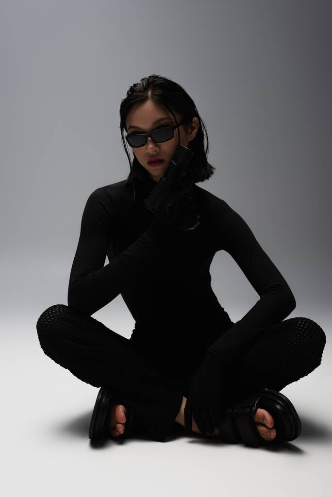 asian woman in total black outfit and stylish sunglasses holding gun while sitting on grey  - Fotoğraf, Görsel