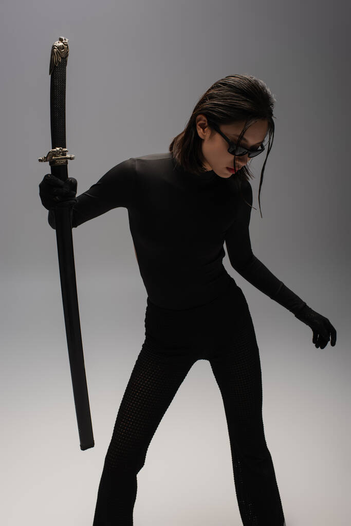 brunette asian woman in black outfit and stylish sunglasses holding katana in scabbard on grey - 写真・画像