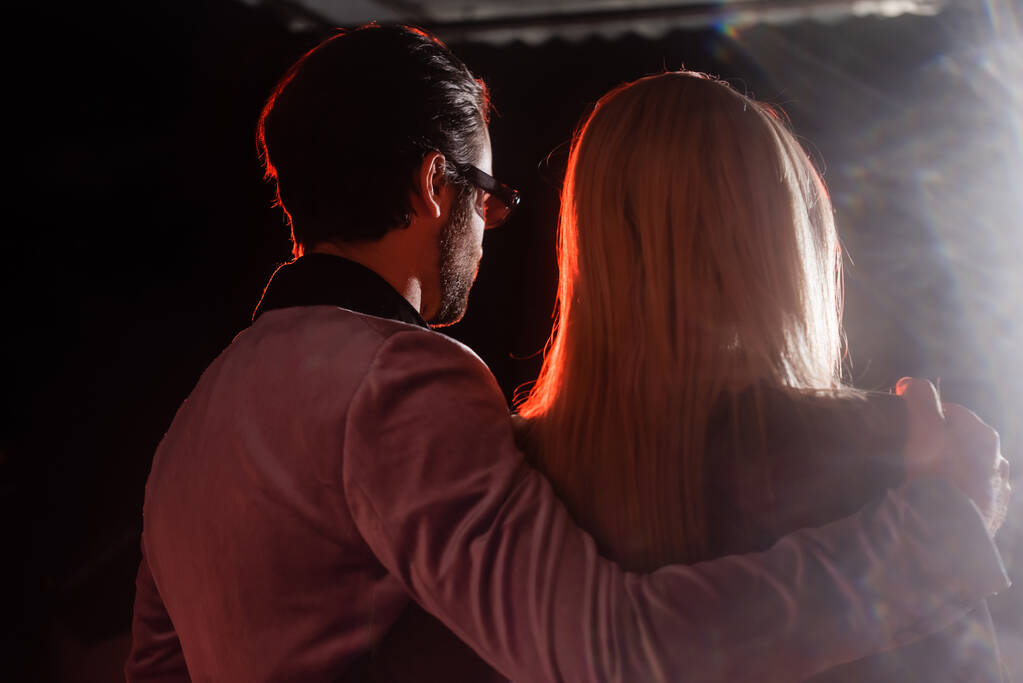 back view of man in trendy blazer and eyeglasses hugging blonde woman on black background with light - Foto, Imagen