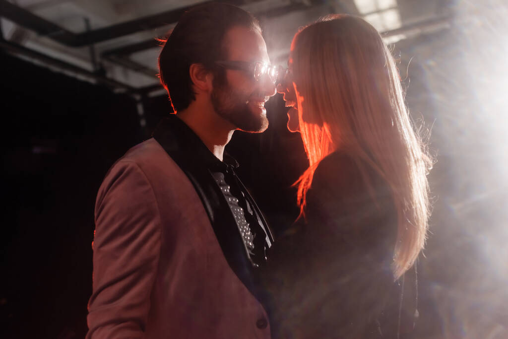 side view of glamour couple in trendy eyeglasses smiling at each other on dark background with light - Fotografie, Obrázek