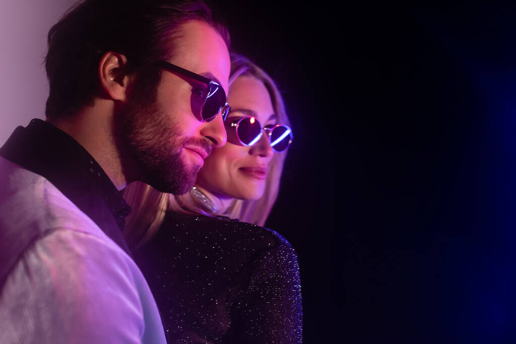 Stylish man in sunglasses looking away near blurred girlfriend during party on purple and black background - Foto, Imagem
