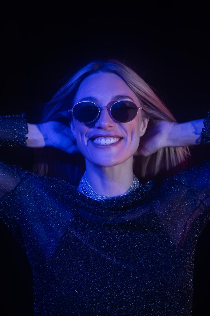 Smiling woman in sunglasses touching hair isolated on black with blue lighting  - Fotografie, Obrázek