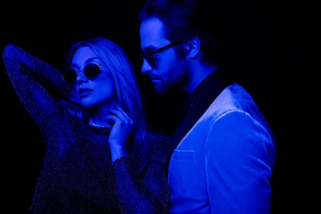 Мужчина в пиджаке и солнцезащитных очках, стоящий рядом со стильной девушкой, изолированный на черном с синим освещением  - Фото, изображение