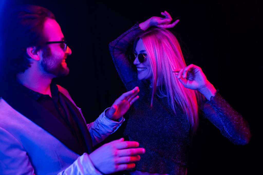 Stylish coupe in sunglasses dancing isolated on black with colorful lighting  - Foto, afbeelding
