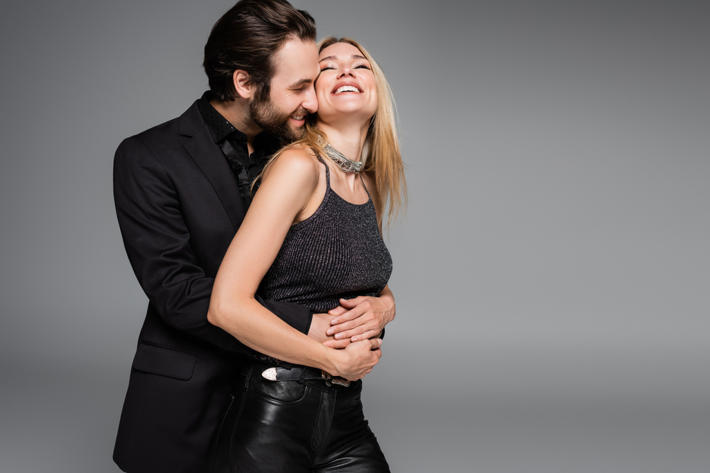 Bearded man in jacket hugging smiling blonde girlfriend isolated on grey  - Photo, Image