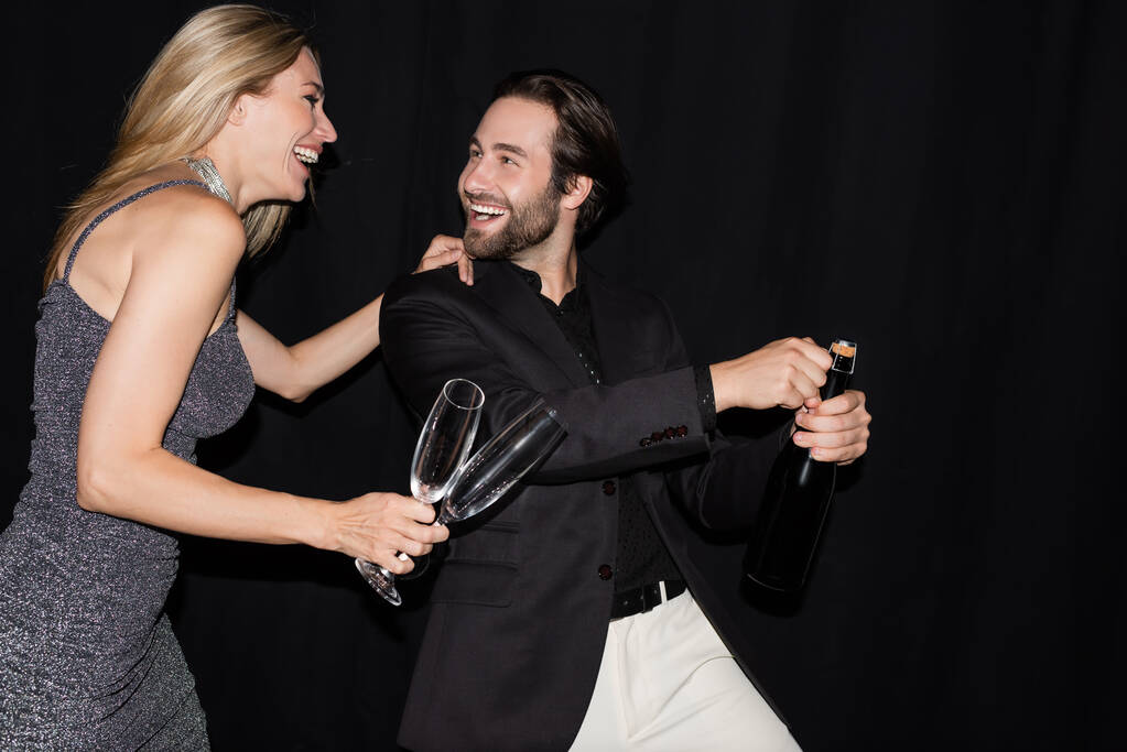 Cheerful woman in dress holding glasses while boyfriend opening champagne isolated on black  - Foto, imagen