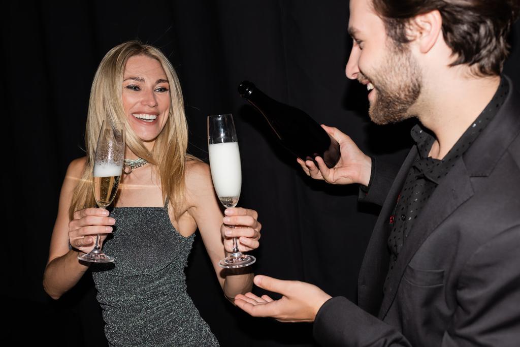 Smiling blonde woman holding glasses while boyfriend pouring champagne isolated on black  - Photo, image