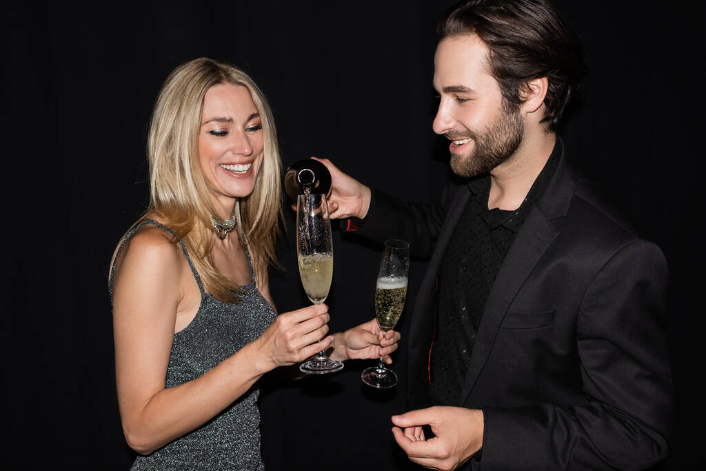 Bearded man pouring champagne near girlfriend in dress isolated on black  - Foto, Imagen