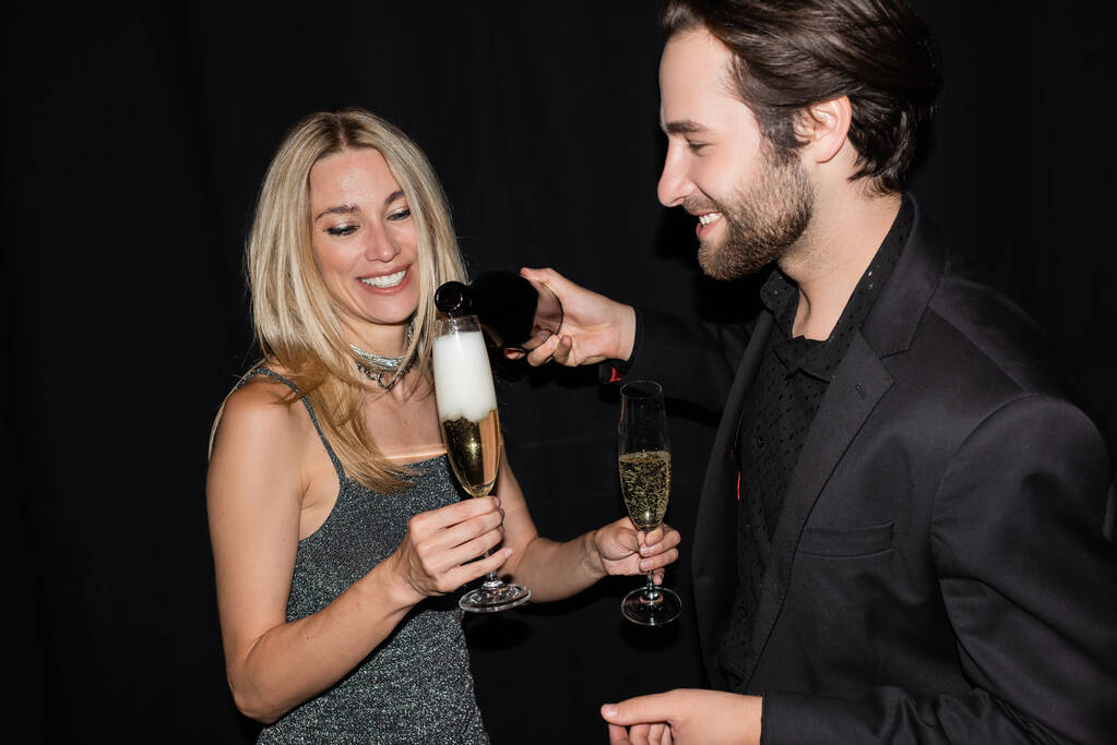 Улыбающийся мужчина в пиджаке разливает шампанское рядом с блондинкой во время вечеринки, изолированной от черных  - Фото, изображение
