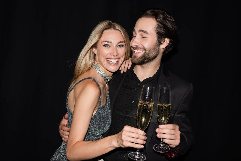 Bearded man hugging cheerful girlfriend with glass of champagne isolated on black  - Photo, image