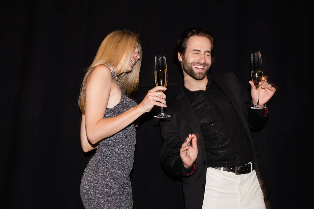 Θετικός άντρας κρατώντας ένα ποτήρι σαμπάνια κοντά στην ξανθιά φιλενάδα του κατά τη διάρκεια πάρτι απομονωμένος σε μαύρο.  - Φωτογραφία, εικόνα