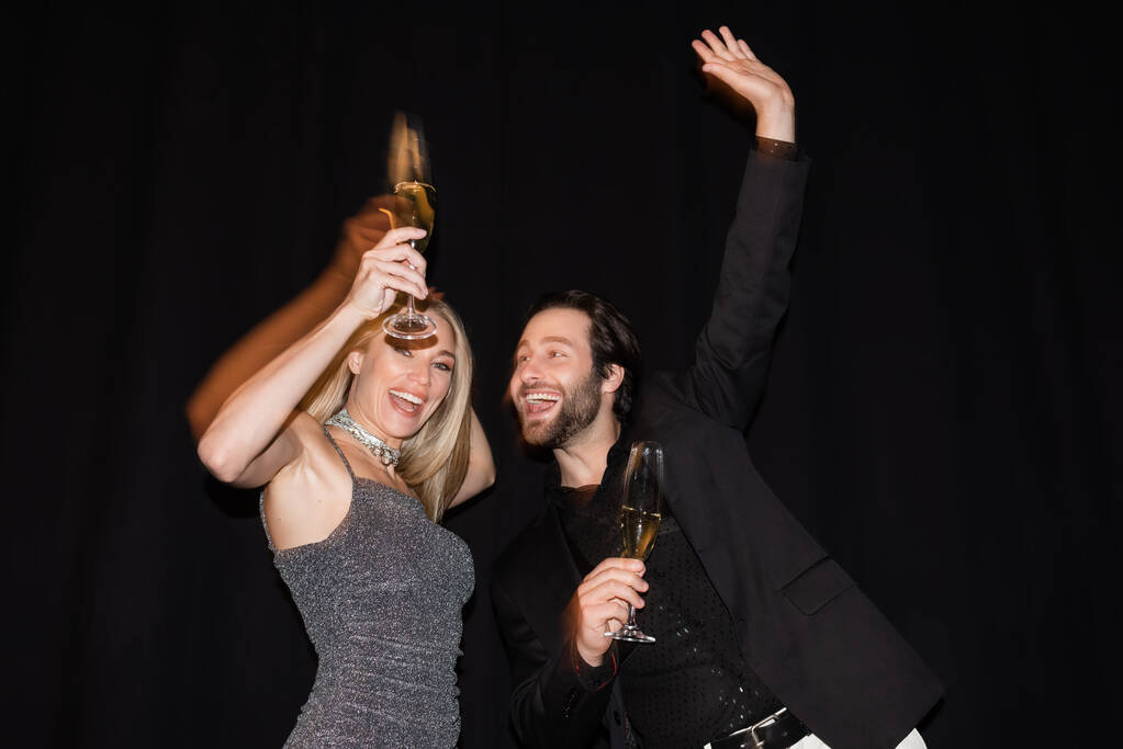 Движение размытой веселой пары, держащей шампанское во время вечеринки изолированы на черном  - Фото, изображение