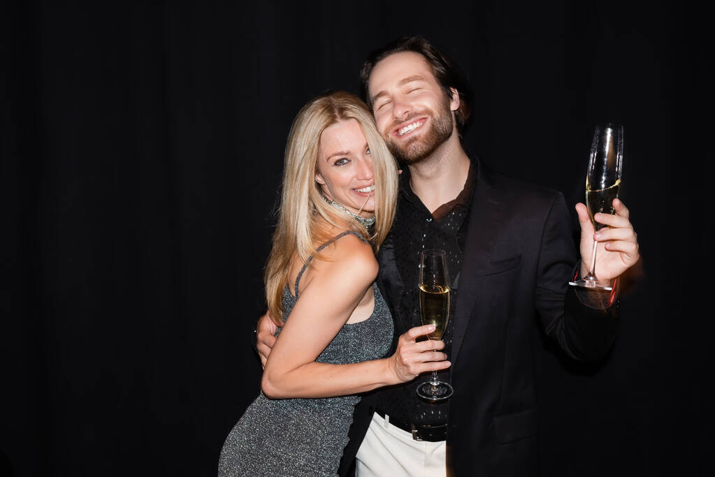 Smiling man holding champagne and hugging sexy girlfriend isolated on black  - Photo, Image