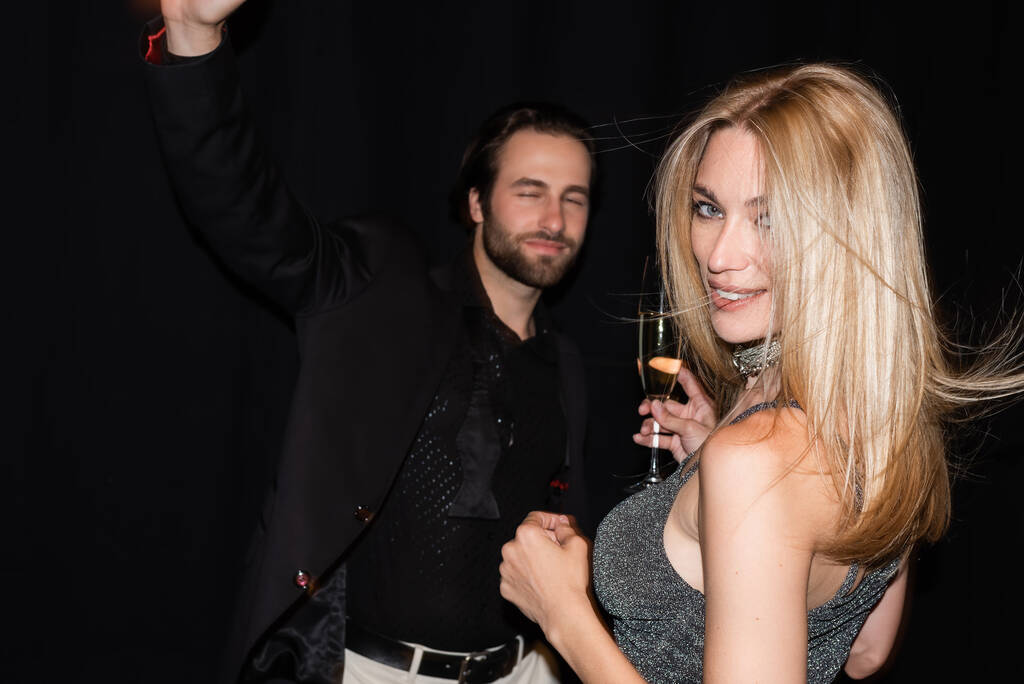 Smiling blonde woman holding glass of champagne while boyfriend dancing isolated on black  - Foto, Bild