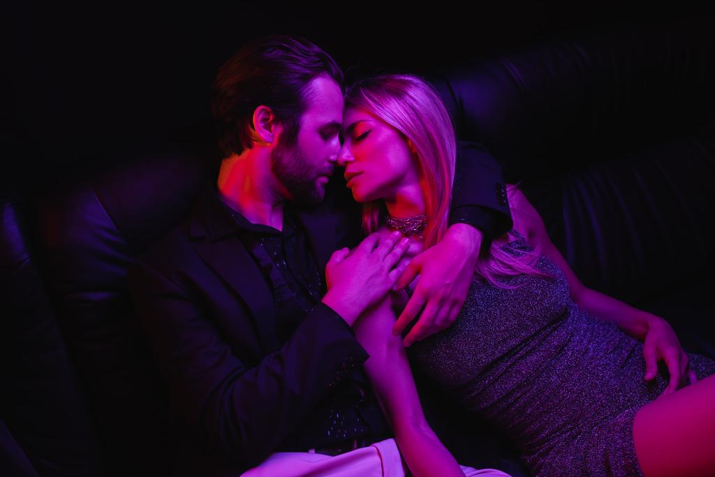 Side view of sexy couple kissing on leather couch with lighting during party isolated on black  - Fotó, kép
