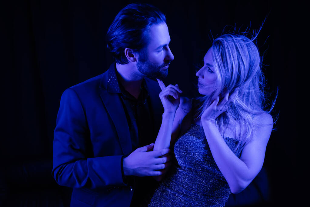 Side view of sexy woman in dress touching boyfriend in jacket with blue lighting isolated on black  - 写真・画像
