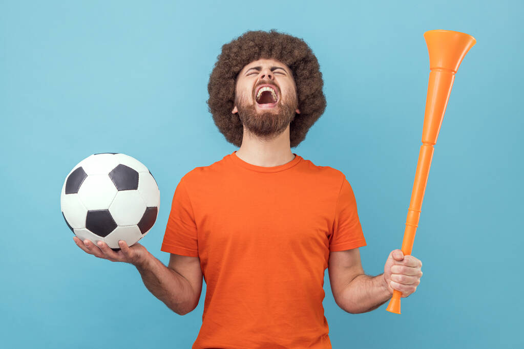 Man met Afro kapsel draagt T-shirt schreeuwend wijdverbreide mond, viert overwinning van favoriete voetbalteam, hoorn en bal in handen. Indoor studio schot geïsoleerd op blauwe achtergrond. - Foto, afbeelding