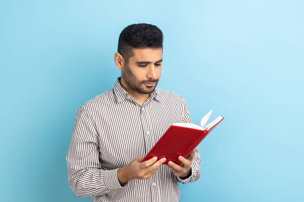 Концентрований бородатий бізнесмен тримає книгу і читання, стоїть поглинений захоплюючим сюжетом, перевіряє графік роботи в блокноті, носить смугасту сорочку. Внутрішній студійний знімок ізольовано на синьому фоні
. - Фото, зображення