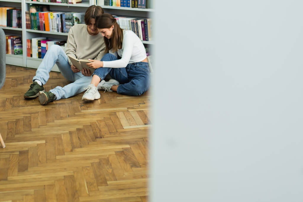 happy teenage couple sitting on floor and reading book near racks in library on blurred foreground - Fotó, kép