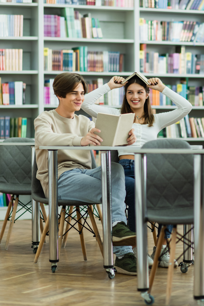 cheerful teen girl covering head with book near smiling friend reading in library - Photo, Image