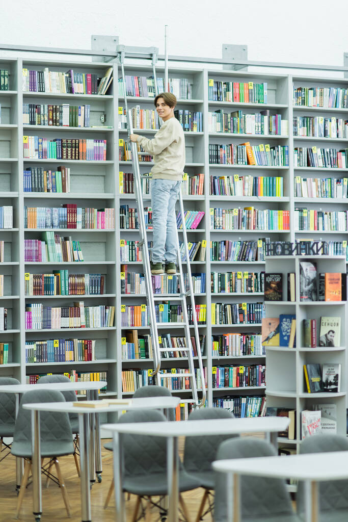 full length of teenage guy smiling at camera from ladder near bookshelves - Fotó, kép