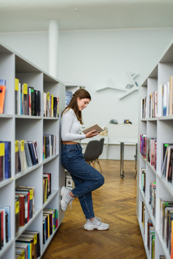 side view of teen girl standing near racks in library and reading book - Photo, Image