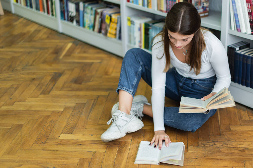 high angle view of teenage girl with books sitting on floor in library - Foto, Imagen