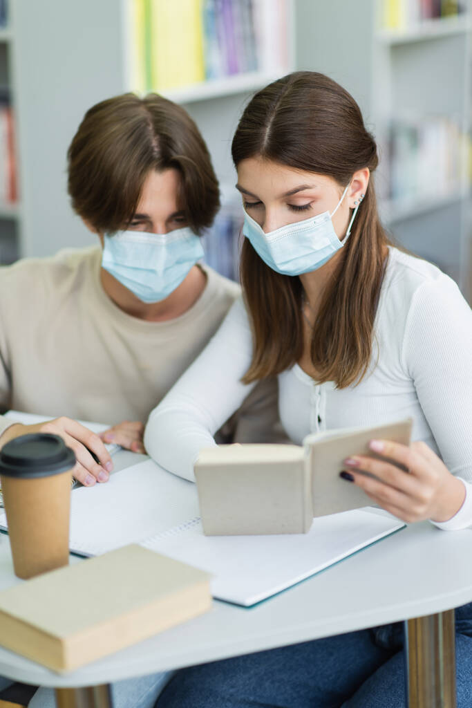 teenage girl in medical mask reading book near paper cup and blurred friend  - Foto, immagini